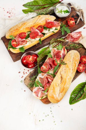 Foto de Two baguette sandwiches with salami, mozzarella cheese, lettuce, tomatoes and ham on a cutting boards. Long subway sandwiches on a white background. Top view. - Imagen libre de derechos