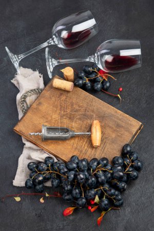 Photo for An overhead photo of glasses of red wine with a bottle, grapes, and a vintage corkscew and corks, shot from above on a dark background. Top view. - Royalty Free Image