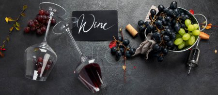 Photo for An overhead photo of glasses of red wine with a bottle, grapes, and a vintage corkscew and corks, shot from above on a dark background. Top view. Panorama. - Royalty Free Image