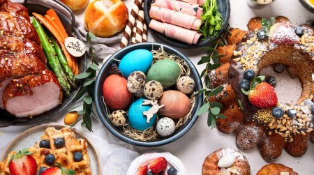 Téléchargez les photos : Traditional Easter dinner or  brunch with ham, colored eggs, hot cross buns, cake and vegetables. Easter meal dishes with holday decorations. Top view - en image libre de droit