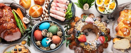 Téléchargez les photos : Traditional Easter dinner or  brunch with ham, colored eggs, hot cross buns, cake and vegetables. Easter meal dishes with holday decorations. Top view, copy space, panorama, banner - en image libre de droit
