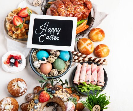 Foto de Traditional Easter dinner or  brunch with ham, colored eggs, hot cross buns, cake and vegetables. Easter meal dishes with holday decorations. Top view, copy space - Imagen libre de derechos