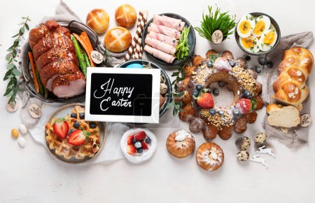 Téléchargez les photos : Traditional Easter dinner or  brunch with ham, colored eggs, hot cross buns, cake and vegetables. Easter meal dishes with holday decorations. Top view, copy space - en image libre de droit