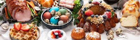 Téléchargez les photos : Traditional Easter dinner or  brunch with ham, colored eggs, hot cross buns, cake and vegetables. Easter meal dishes with holday decorations. Panorama, banner - en image libre de droit