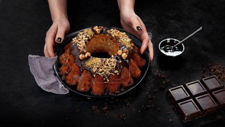 Foto de Bundt chocolate cake decorated with melted chocolate, nuts  and sprinkles on plate with woman hand on black background. Holiday concept. - Imagen libre de derechos