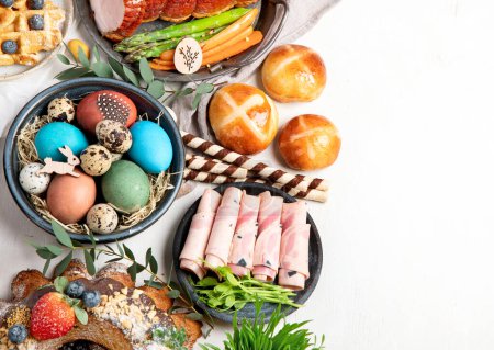 Photo for Traditional Easter dinner or  brunch with ham, colored eggs, hot cross buns, cake and vegetables. Easter meal dishes with holday decorations. Top view, copy space - Royalty Free Image