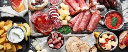 Photo for Typical spanish tapas with jamon slices, chorizo, salami,  olives,  potatoes snack Patatas bravas, seafoods on dark table. Traditional spanish food. Top view, panorama, banner - Royalty Free Image