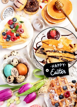 Foto de Easter sweet dessert table. Pancakes, crepes, waffles and donuts  with fresh berries, nuts and topping.   Easter traditional natural colorful eggs. Top view - Imagen libre de derechos