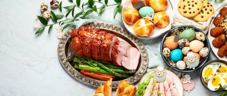 Photo for Classic Easter ham dinner. Top view table scene on a white background. Ham, eggs, hot cross buns, carrot, cake and vegetables. Panorama with copy space. - Royalty Free Image