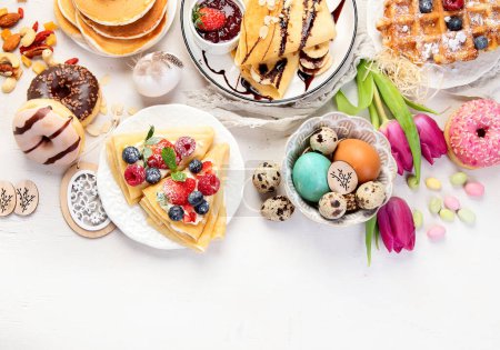 Foto de Easter sweet dessert table. Pancakes, crepes, waffles and donuts  with fresh berries, nuts and topping.   Easter traditional natural colorful eggs. Top view, copy space - Imagen libre de derechos