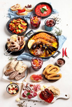 Foto de Typical spanish tapas concept. Concept include jamon, chorizo sausage, brushettas, bowl with olives, shrimp, pan with paella, cheese, sangria, churros on a white background. Top view. - Imagen libre de derechos