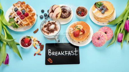 Foto de Summer or spring breakfast with Cup of hot cappuccino coffee,  pancakes, waffles coffee, tulips  flowers, craft envelope on blue background.  Top view, flat lay - Imagen libre de derechos
