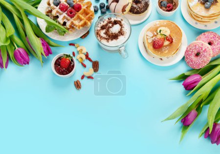 Foto de Summer or spring breakfast with Cup of hot cappuccino coffee,  pancakes, waffles coffee, tulips  flowers, craft envelope on blue background.  Top view, flat lay, copy space - Imagen libre de derechos