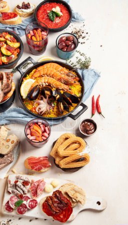 Photo for Typical spanish tapas concept. Concept include jamon, chorizo sausage, brushettas, bowl with olives, shrimp, pan with paella, cheese, sangria, churros on a white background. Top view. Copy space. - Royalty Free Image