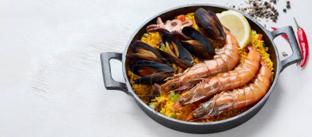 Photo for Classic dish of Spain, seafood paella in traditional pan on a white background, top view. Spanish paella with shrimps, clamps, mussels, green peas and fresh lemon wedges from above. Panorama with copy space. - Royalty Free Image