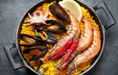 Photo for Classic dish of Spain, seafood paella in traditional pan on a dark background, top view. Spanish paella with shrimps, clamps, mussels, green peas and fresh lemon wedges from above. - Royalty Free Image