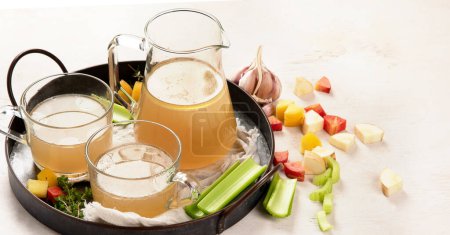 Photo for Two cups and glass mug of homemade chicken bone broth on white background. Bone broth, bone stock, garlic cloves and herbs. Panorama. - Royalty Free Image