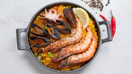 Photo for Classic dish of Spain, seafood paella in traditional pan on a white background, top view. Spanish paella with shrimps, clamps, mussels, green peas and fresh lemon wedges from above. - Royalty Free Image