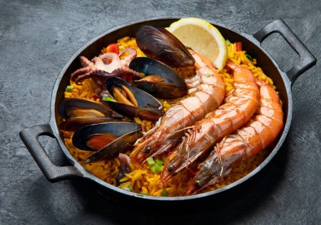 Photo for Classic dish of Spain, seafood paella in traditional pan on a dark background, top view. Spanish paella with shrimps, clamps, mussels, green peas and fresh lemon wedges from above. - Royalty Free Image