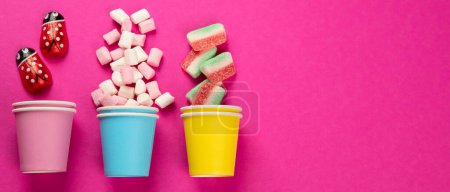 Téléchargez les photos : Assortiment de bonbons à la gelée douce multicolore et de guimauves dans des tasses éco sur fond rose. Vue de dessus. Panorama avec espace de copie. - en image libre de droit