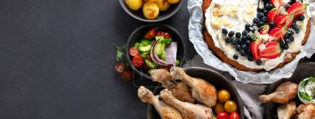 Photo for Dinner with chicken, vegetables, cake, salad and fruits on a dark background. Summer food concept. Top view. Panorama with copy space. - Royalty Free Image