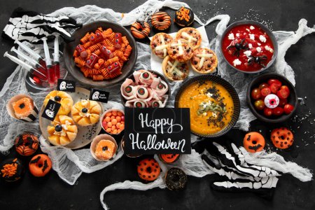Photo for Fun Halloween dinner party table. Pizza, pumpkin soup, candy, eyeball spaghetti, snacks and donuts on a black background. Top view. - Royalty Free Image