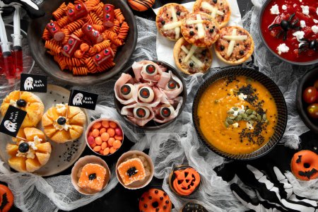 Photo for Fun Halloween dinner party table. Pizza, pumpkin soup, candy, eyeball spaghetti, snacks and donuts on a black background. Top view. - Royalty Free Image