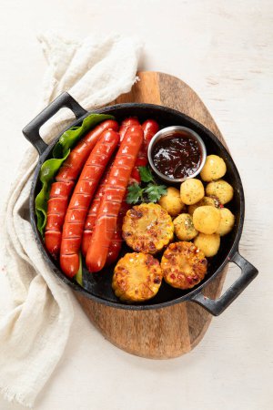 Téléchargez les photos : Saucisses grillées avec pommes de terre et maïs dans une casserole sur fond blanc. Vue du dessus. - en image libre de droit