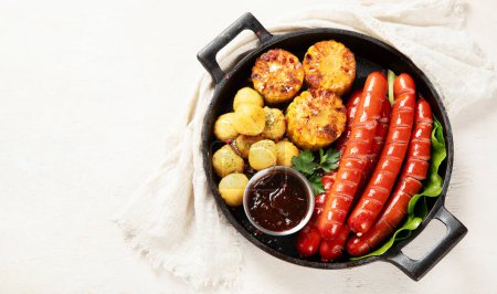Téléchargez les photos : Saucisses grillées avec pommes de terre et maïs dans une casserole sur fond blanc. Vue de dessus. Espace de copie. - en image libre de droit