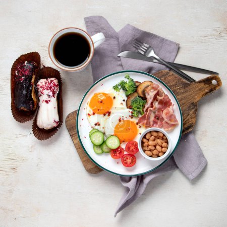 Foto de Plato de desayuno tradicional inglés con tiras de tocino, huevos soleados, verduras y pastel sobre fondo claro. - Imagen libre de derechos