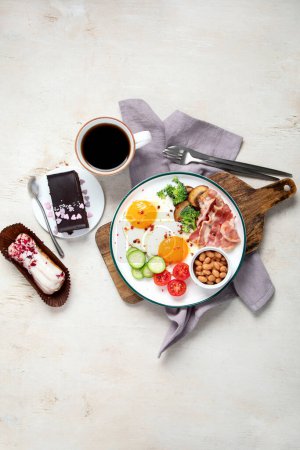 Photo for Traditional Englis breakfast plate with bacon strips, sunny side up eggs, vegetables and cake on light background. - Royalty Free Image