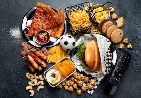 Photo for Saturated fats. Football time. TV remote control and snacks - chips, popcorn, cookies, cheese, sauce, fries, burger, nuts. Top view - Royalty Free Image