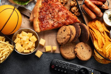Photo for Unhealthy food i basket. TV remote control and snacks - chips, popcorn, cookies, cheese, sauce, fries, pizza. Top view - Royalty Free Image