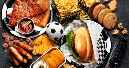 Photo for Saturated fats. Football time. TV remote control and snacks - chips, popcorn, cookies, cheese, sauce, fries, burger, nuts. Top view - Royalty Free Image