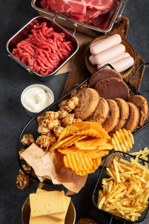 Photo for Saturated and trans. Unhealthy food. Sausage, potato, meat, cheese, popcorn, cookies, cream on dark background. - Royalty Free Image