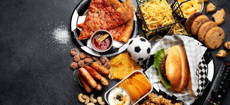 Photo for Saturated fats. Football time. TV remote control and snacks - chips, popcorn, cookies, cheese, sauce, fries, burger, nuts. Top view, banner, copy sapce. - Royalty Free Image