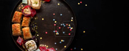 Téléchargez les photos : Ramadan nourriture sur une grande assiette. Bonbons arabes - lokum, confiture de fruits, baklava sur fond noir. Vue du dessus, espace de copie, bannière. - en image libre de droit