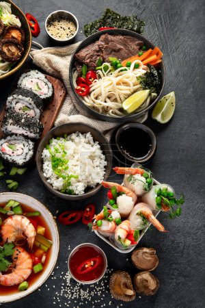 Foto de Mesa de comida tradicional asiática. Rollo de primavera, arroz, camarones, sushi, verduras, carne sobre fondo oscuro. Vista superior - Imagen libre de derechos