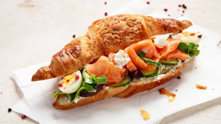 Foto de Desayuno saludable salado sobre fondo claro. Croissant de salmón fresco con queso crema, pepino, huevo y ensalada. - Imagen libre de derechos