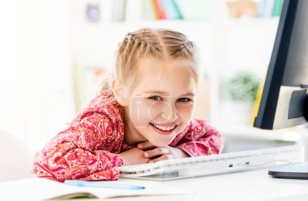 Photo for Smiling little girl sitting at computer desk in bright room - Royalty Free Image