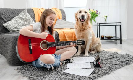 Foto de Chica adolescente practicando guitarra jugando con perro golden retriever en casa sentado en el suelo. Guitarrista bastante con instrumento músico y perrito de raza pura mirando a la cámara - Imagen libre de derechos