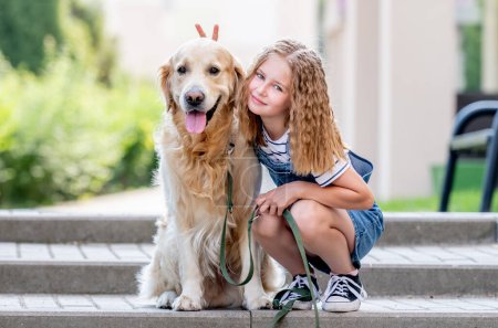 Photo for Preteen girl wearing hat hugging golden retriever dog sitting outdoors in summertime. Pretty kid petting fluffy doggy pet in city - Royalty Free Image