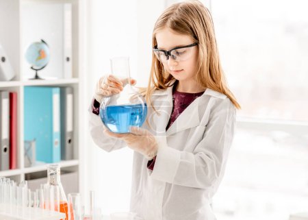 Foto de Chica inteligente haciendo experimento de química científica usando gafas de protección, sosteniendo la botella y midiendo líquido azul. Colegiala con equipo químico en la lección escolar - Imagen libre de derechos