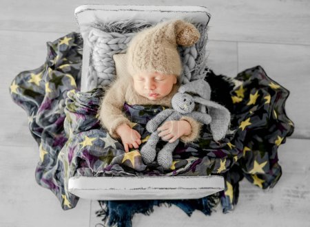 Téléchargez les photos : Nouveau-né garçon dormant sur la fourrure dans un petit lit. Adorable enfant en bas âge qui fait une sieste sur son ventre pendant la séance photo en studio portant un chapeau et un costume tricotés - en image libre de droit
