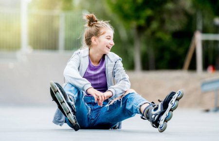 Foto de Cute girl roller skater sitting on ground in city park and smiling. Pretty female teenager with hairstyle posing during rollerskating - Imagen libre de derechos