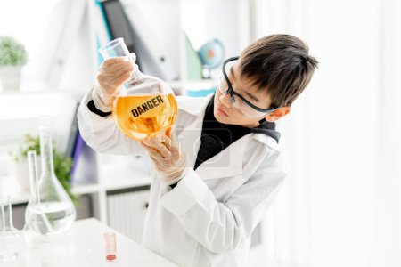 Téléchargez les photos : Un écolier portant des lunettes de protection faisant des expériences de chimie en classe de sciences élémentaires. Tube de maintien de la pupille intelligent avec marque de danger en laboratoire pendant l'essai - en image libre de droit