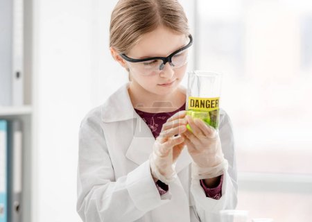 Photo for Smart girl doing scientific chemistry experiment wearing protection glasses, holding bottle with title danger. Schoolgirl with chemical equipment on school lesson - Royalty Free Image