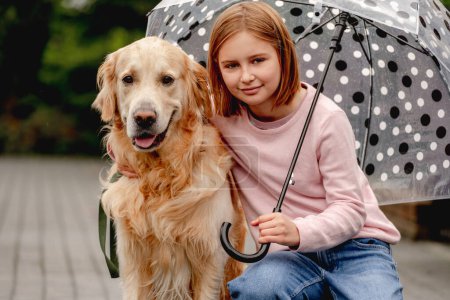 Foto de Chica preadolescente con perro golden retriever sentado bajo el paraguas en la calle y mirando hacia atrás. Niño bonito con mascota perrito en día lluvioso - Imagen libre de derechos