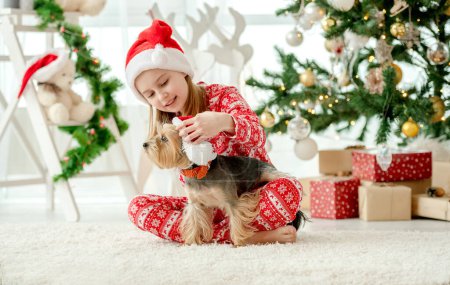 Foto de Niño niña acariciando perro con sombrero de Santa en casa en época de Navidad. Niño hembra con mascota perrito en Año Nuevo - Imagen libre de derechos
