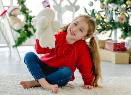 Foto de Niña con juguete de conejito en Navidad sentada en la alfombra en la habitación con árbol decorado y regalos. Niño bonito en casa en vacaciones de Año Nuevo - Imagen libre de derechos